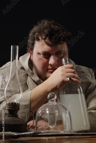 A fat, warlike monk at a table on which stands a bottle of elixir or simply moonshine, a kerosene lamp, some kind of magical utensils and a large two-handed sword