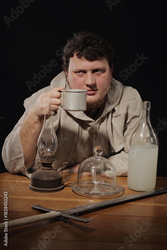 A fat, warlike monk at a table on which stands a bottle of elixir or simply moonshine, a kerosene lamp, some kind of magical utensils and a large two-handed sword