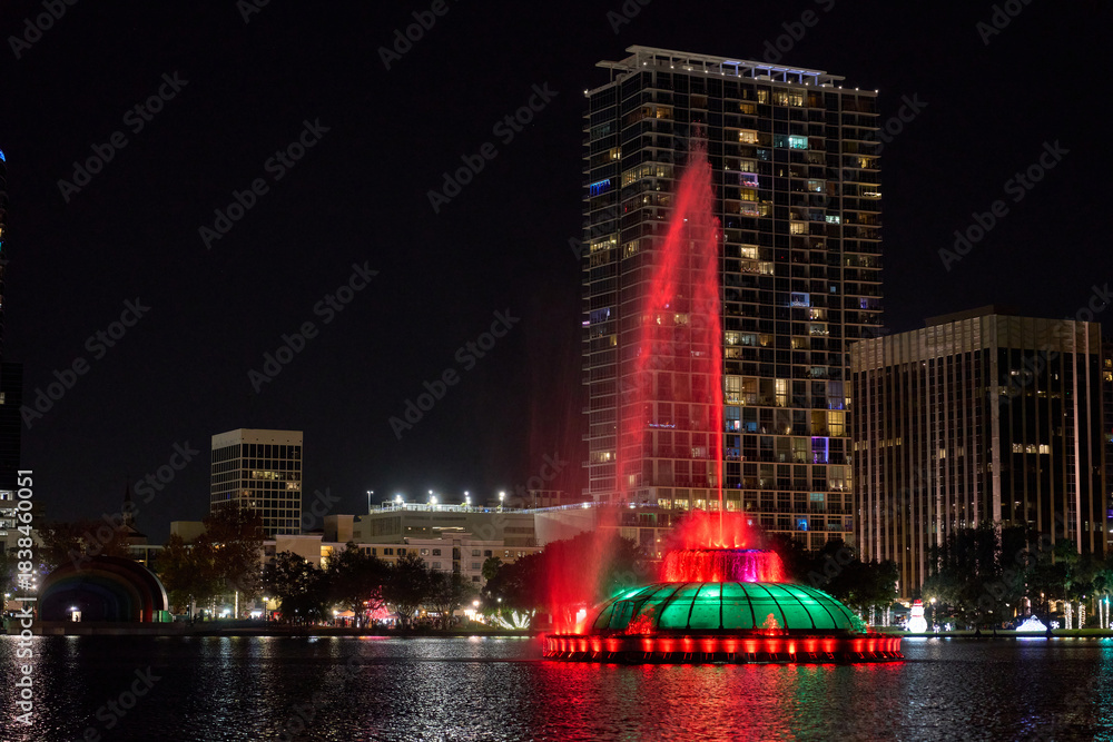 Obraz premium A tall water fountain glows red and green on the lake against modern high-rises. Reflections shimmer across the downtown cityscape.