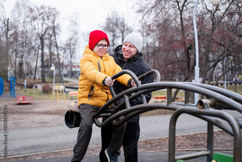 A 40-year-old man in a warm jacket and hat pushes his son, wearing a yellow jacket, on a metal swing in a park. A happy boy and his father on a swing.