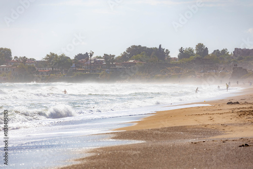 Fototapeta Naklejka Na Ścianę i Meble -  Powerful waves crashing on a sunlit beach in dramatic backlight, creating a misty spray with silhouettes of people. A dynamic scene on the Mediterranean coast. Side, Antalya, Turkey.


