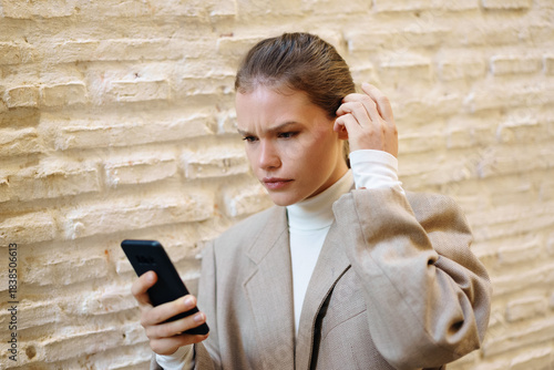 A young woman stands by a textured wall, using her smartphone and showcasing her style