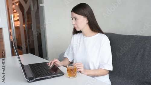 young beautiful caucasian brunette woman freelancer works in cafe with notebook, casual clothes, white T-shirt, glass with apple juice, grey couch and notebook
