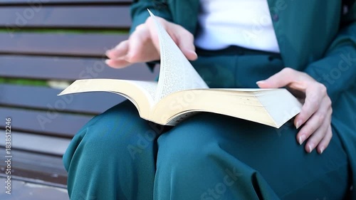 young woman holds thick book in hands, reading book, sitting on bench in park, close-up view, no face