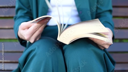 young woman holds thick book in hands, reading book, sitting on bench in park, close-up view, no face