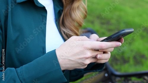 young woman holds smartphone in hands, texting, watching content, sitting on bench in park, close-up view, no face, copy space