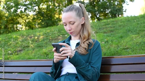 young woman holds smartphone in hands, texting, watching content, sitting on bench in park