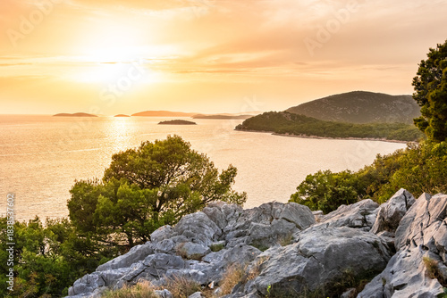 An orange sunset over the sea with a view of small islands. The sun is low on the horizon behind thick clouds, and the coastal landscape is visible from a mountaintop vantage point.