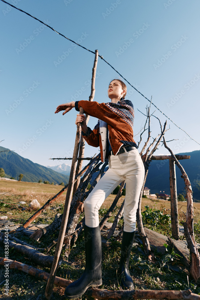 Fototapeta premium Woman outdoors in mountains leaning on a wooden structure in a rural field, wearing textured sweater, white pants and black boots, portrait with clear sky, sunlight and rustic mood.