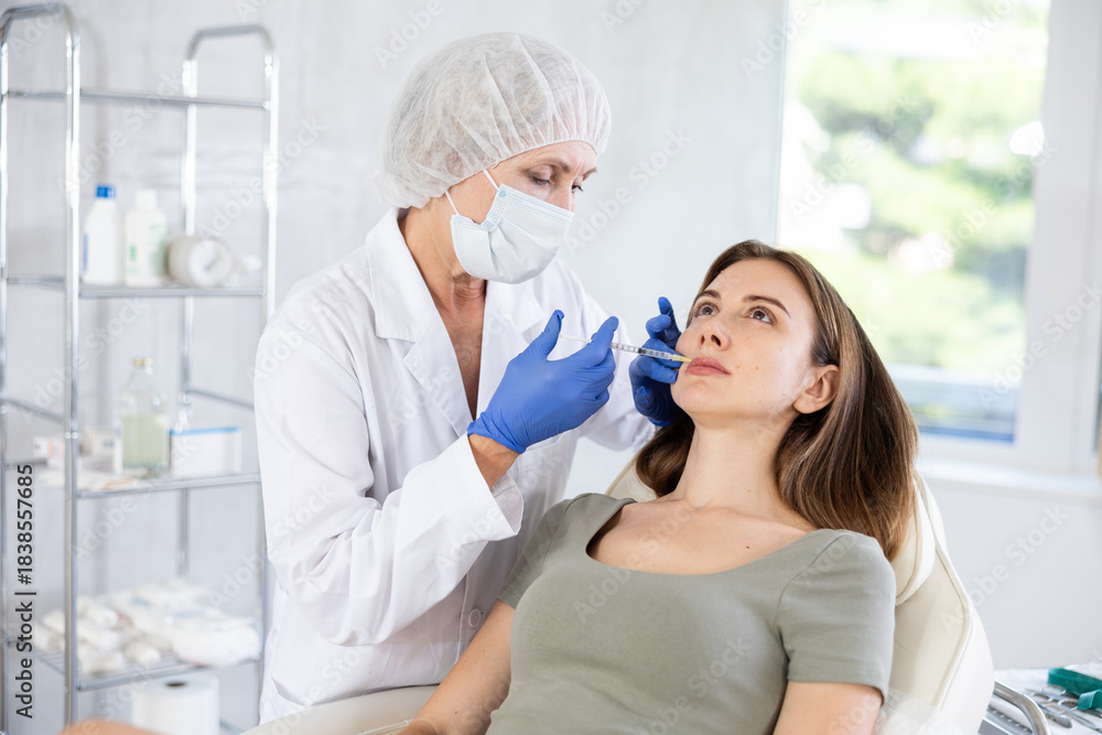 Obraz premium Doctor wearing uniform, medical mask and gloves giving beauty injection to young female patient at treatment room of beauty clinic