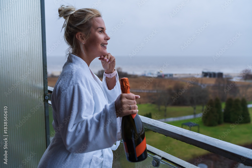 Fototapeta premium Happy Young Woman in White Bathrobe Holding Bottle of Sparkling Wine on Hotel Balcony Overlooking Beach in Parnu Estonia.
