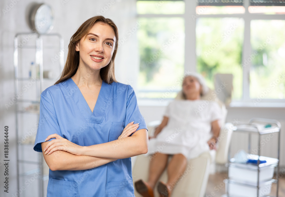 Fototapeta premium Portrait of young female doctor in scrub standing while senior patient laying in the back.