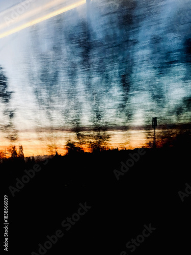 sunset over the city, Abstract blurred silhouettes of trees and deep shadows contrast with vibrant streaks of blue, orange, and golden sky colors