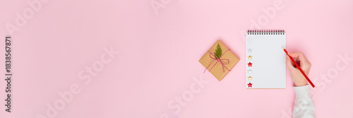 Hand writing on blank notepad next to wrapped gift box on pastel pink background with wide copy space for holiday banners, wish lists, new year planning, marketing layouts and festive design projects.