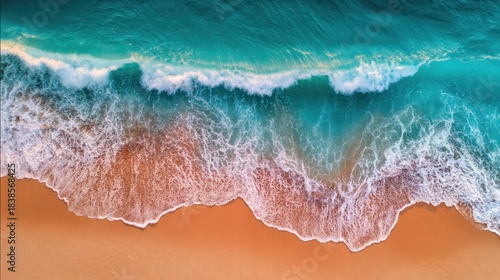 Waves crash onto a sandy beach creating foam as clear blue water moves towards the shore. The scene captures the ocean's movement and sandy texture under sunlight.