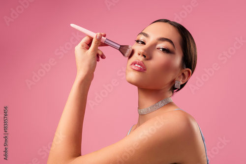 Gorgeous young woman applying makeup with a brush against a soft pink background, showcasing graceful beauty