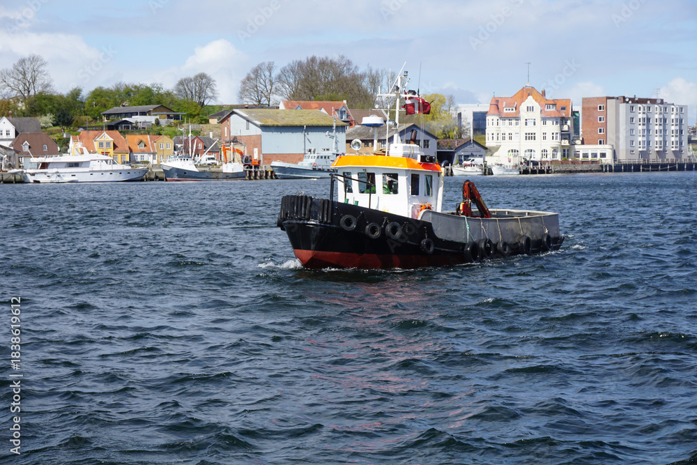 Fototapeta premium Small tugboat navigating dark blue choppy waters in Sønderborg Harbor, Denmark, with colorful waterfront buildings in the background