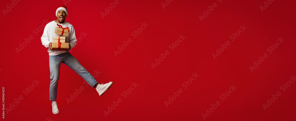 Fototapeta premium A man wearing a Santa hat joyfully jumps in the air while holding wrapped gifts. The vibrant red background enhances the festive mood. It's a lively celebration of the holiday spirit.