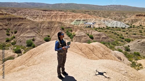 Woman operating drone in vast desert landscape with rugged terrain under clear sky.