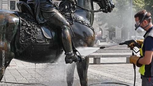 Worker cleaning a bronze statue with a pressure washer in a public square.