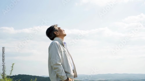 Elderly man standing hillside looking sky with serene profile, peaceful mountain landscape, jacket clad contemplative figure bathed soft sunlight