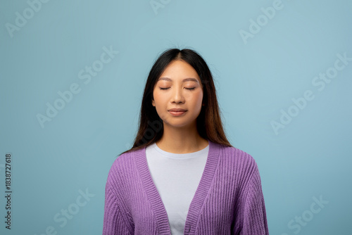A young woman wearing a purple cardigan stands still with her eyes closed, embracing a moment of mindfulness. The light blue background creates a calming atmosphere.