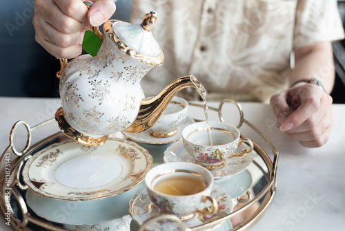 Close-up of a person pouring hot tea from a vintage floral porcelain teapot into a matching cup. Served on a golden tray for afternoon tea time.