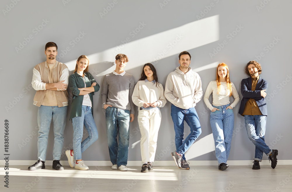 Obraz premium Group of fashion models posing in casual clothing. Seven happy young people wearing comfortable informal clothing standing by a gray wall in sunlight. Fashion concept