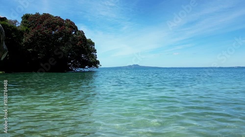 Low drone aerial moving out from Winstones Cove toward clear water with Rangitoto beyond in New Zealand.
