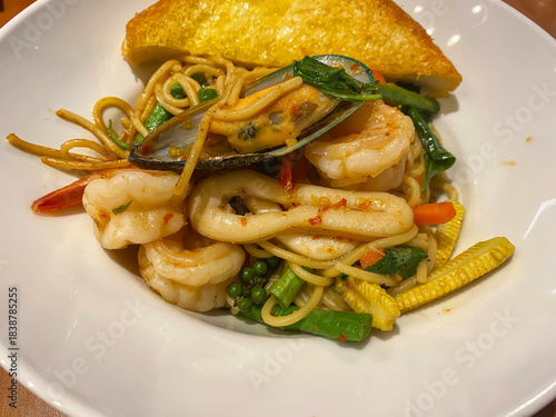 Spicy seafood spaghetti stir-fry featuring shrimp, squid, mussel, and herbs, served with a crispy slice of garlic bread on a white plate.