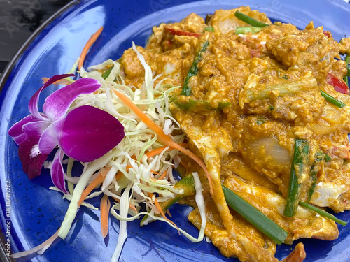 A rich and creamy stir-fried crab dish with yellow curry powder, egg, and vegetables, garnished with a purple orchid on a blue plate.