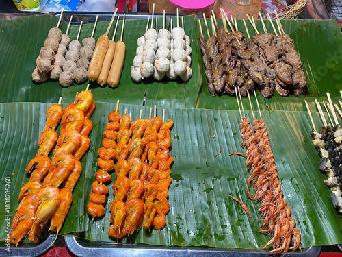 A variety of Thai street food skewers including meatballs, sausages, grilled organ meat, and small shrimps displayed on fresh green banana leaves.