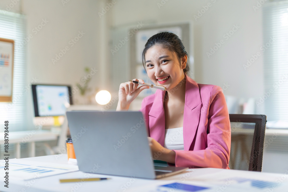 Obraz premium Young Asian professional woman in a pink blazer smiling confidently while using a laptop at her desk, working efficiently