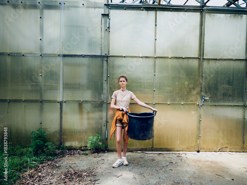 Cuadro en lienzo Woman in front of greenhouse holding a large black bucket, casual clothing and sneakers, gardening scene with plants and soil, outdoor agriculture and horticulture activity