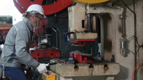 Senior industrial worker with grey beard and safety gloves operating a heavy red hydraulic press. He processes metal parts in a manufacturing factory workshop.