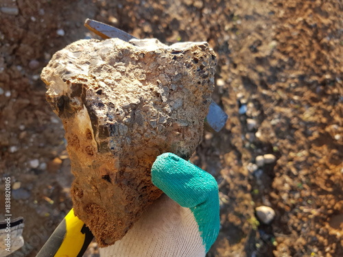 Rough, earthy flint concretion held in a gloved hand against a blurred background of dry soil and small stones, showcasing geological texture and color contrast