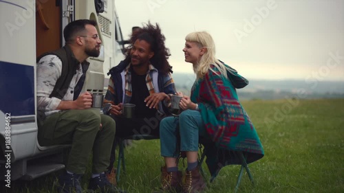 Happy multiracial friends sitting on camping chairs next to their recreational vehicle, laughing and talking while enjoying a warm beverage in a beautiful natural landscape setting