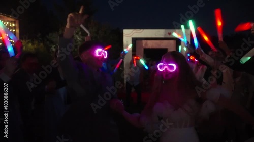 On a wedding party newlywed couple in glowing glasses walks through a crowd of guests holding colorful LED sticks at night. Diverse group of friends and relatives. Translation: Alex and Stacy