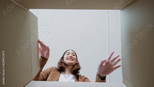 Low angle view of surprised young asian woman unpacking. Opening carton box and looking inside. Packaging box, delivery service. Human emotions and facial expression