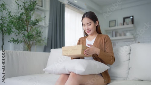 Happy young Asian woman opening delivery package at home. Beautiful female customer open box and sitting on couch in living room