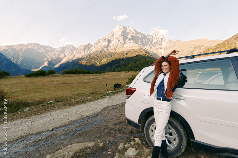 Fototapeta premium Woman by car in mountain landscape, smiling with arms raised beside white SUV on a gravel road, travel adventure in an open meadow with scenic peaks, clear sky and relaxed vibe.