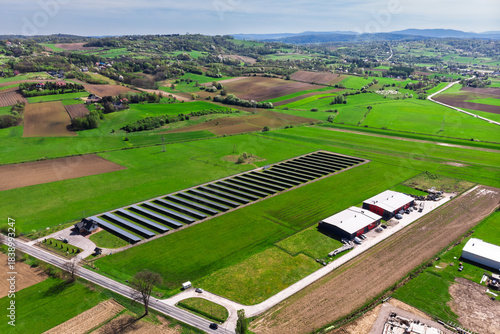 Renewable energy source. Aerial panorama of a massive solar PV power plant installation surrounded by lush green fields in a rural setting, symbolizing technology and sustainability.