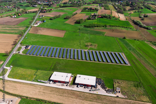 Renewable energy source. Aerial panorama of a massive solar PV power plant installation surrounded by lush green fields in a rural setting, symbolizing technology and sustainability.