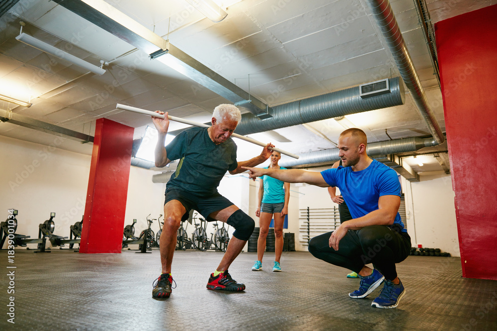Fototapeta premium Coach, old man or squat in gym class with pipe for mobility, fitness activity or exercise instructions. Personal trainer, senior person or pvc equipment with help for workout, stability or low angle.