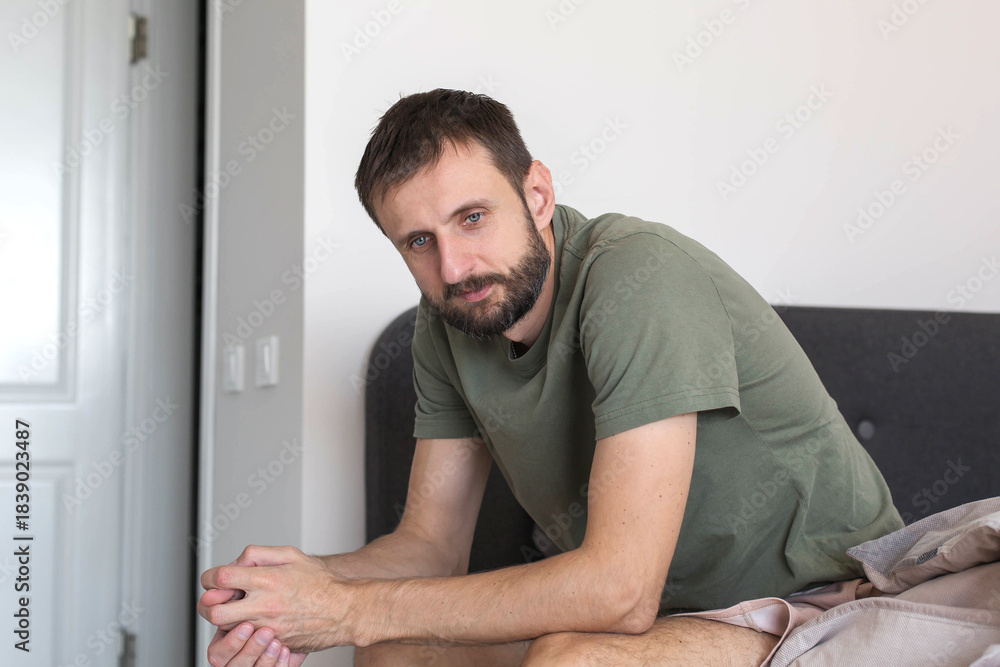 Fototapeta premium A tired man sitting on the edge of a bed in a bright modern bedroom, leaning forward with clasped hands and a thoughtful morning expression, showing a realistic everyday waking moment