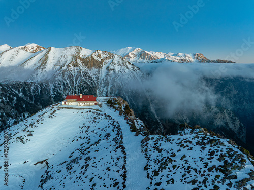 Aerial view of a snow-laden mountain peak capped by a charming red-roofed building, amidst the rugged, snow-dusted landscape, Omalos, Chania, Greece.