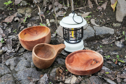 A white portable hanging electric camping lantern with wooden plates, bowls, and mugs placed on soil and rocky ground during outdoor activities.