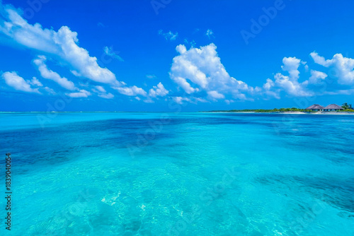 Bright blue ocean waters and clear sky fill the view from a tropical beach. Clouds drift over the horizon in the distance