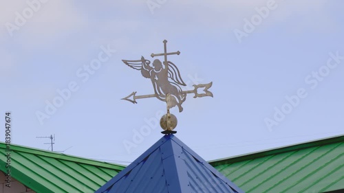 Metal weather vane with angel and cross silhouette mounted on blue roof peak, green metal roofs and pale blue sky in background.