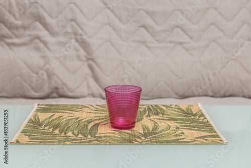 Pink glass cup on a palm leaf placemat on a table with a beige background